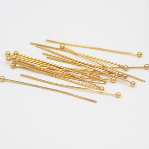 May include: A group of gold-colored head pins with round heads. The pins are scattered on a white surface.