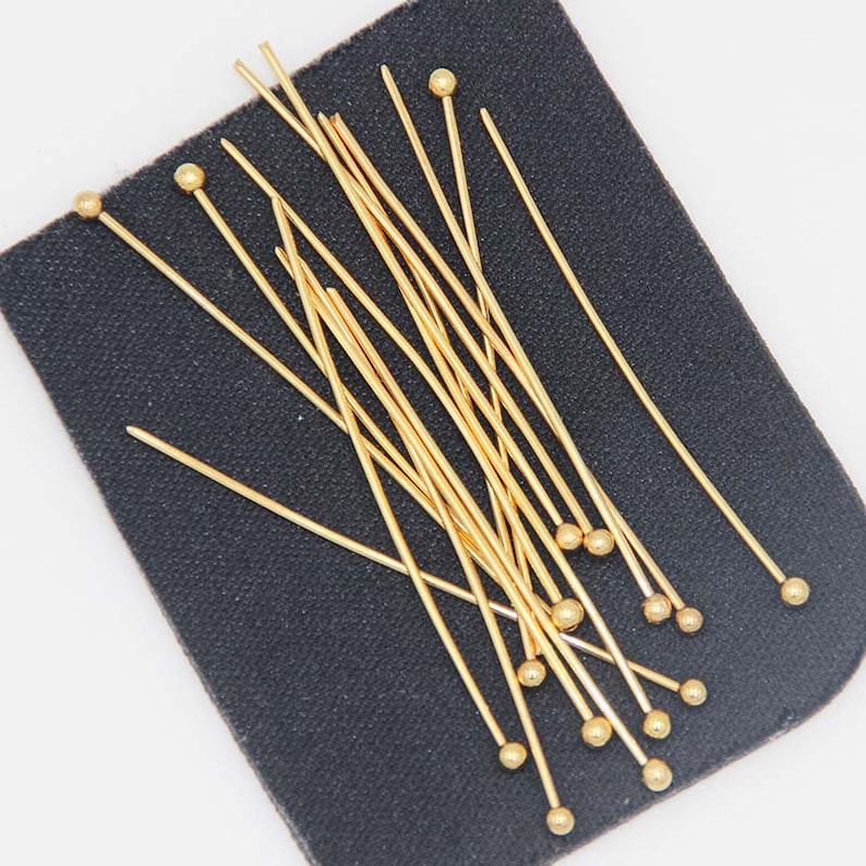 May include: A group of gold-colored head pins with round heads. The pins are arranged on a black surface.