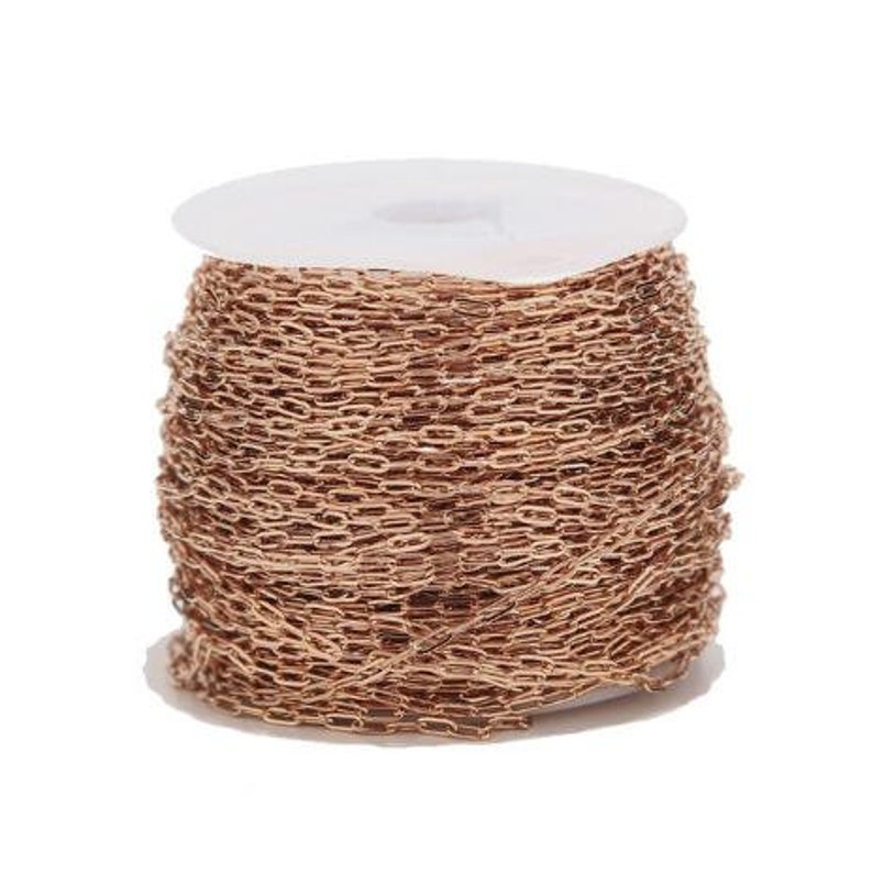 Op de afbeelding: Een spoel met ros&eacute;goudkleurige ketting. De ketting bestaat uit kleine, ovaalvormige schakels en is strak gewikkeld rond een witte kartonnen spoel. De ketting heeft een warme, metallic kleur en lijkt nieuw.