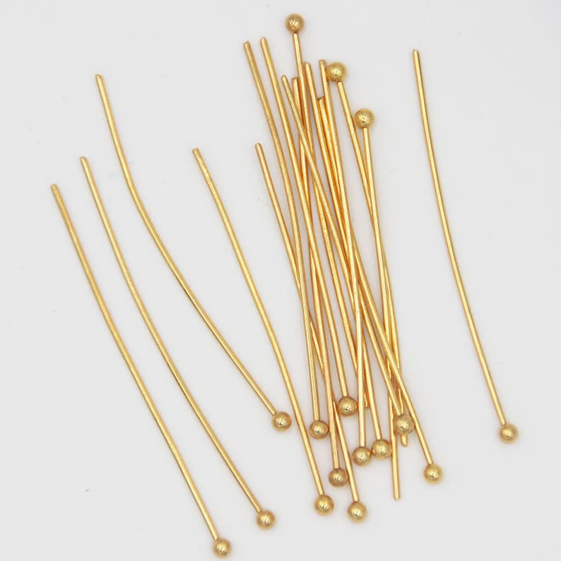 May include: A group of gold head pins with round heads. The pins are arranged in a random pattern on a white background.