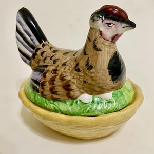 May include: A ceramic hen figurine with a brown and black body, a red head, and a black beak. The hen is sitting on a green grassy nest with white eggs, inside a yellow basket-shaped base. Decorative kitchenware.