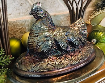 May include: A iridescent purple glass hen with a detailed design sits on a silver platter. The hen is perched on a bed of leaves and has a detailed design on its feathers.