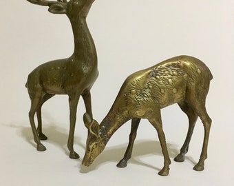 May include: Two brass deer figurines. One deer stands upright with antlers, head raised. The other deer is in a grazing position, head lowered. Both have detailed features and a metallic finish, suitable for home decor.