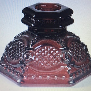 May include: A dark red glass candle holder with a hexagonal base and a decorative design. The holder has a raised, octagonal top with a central opening for a candle.