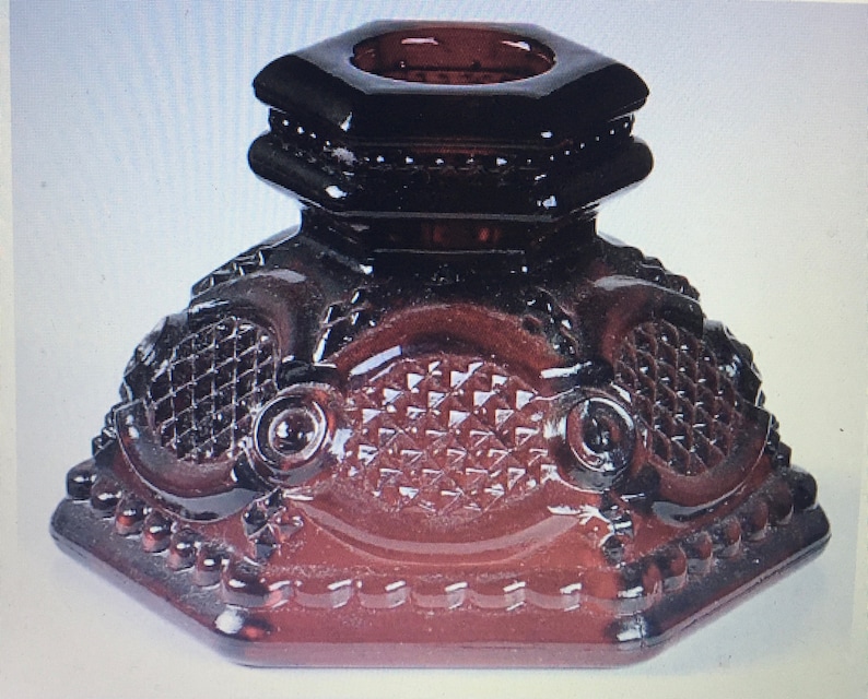 May include: A dark red glass candle holder with a hexagonal base and a decorative design. The holder has a raised, octagonal top with a central opening for a candle.