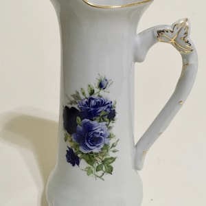 May include: White porcelain pitcher with a gold trim and a blue rose floral design. The pitcher has a tall, slender body and a curved handle with a leaf design.