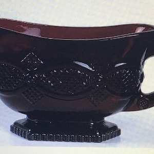 May include: A dark burgundy glass gravy boat with a decorative diamond pattern. The boat has a handle and a footed base.