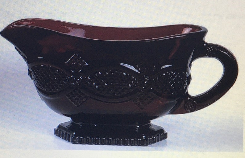 May include: A dark burgundy glass gravy boat with a decorative diamond pattern. The boat has a handle and a footed base.