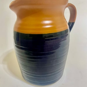 Puede incluir: Una jarra de cerámica marrón y negra con una boca ancha y un asa. La jarra tiene una parte superior marrón y una parte inferior negra con un sutil patrón de nervaduras.