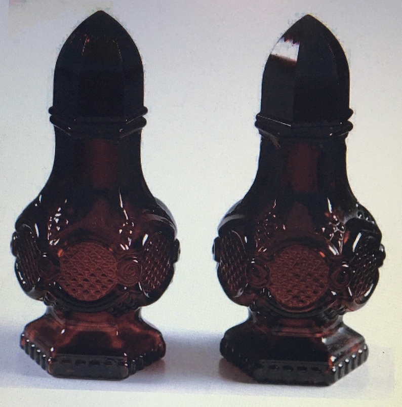 May include: A pair of dark red glass salt and pepper shakers with intricate designs. The shakers have a rounded top and a wide base.
