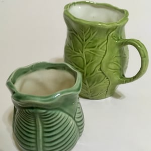 May include: Two green ceramic mugs with leaf designs. The larger mug has a handle and the smaller mug is a planter.