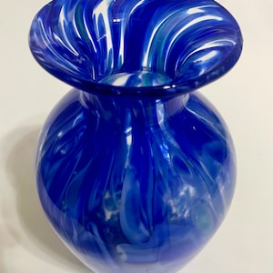 Puede incluir: Un jarrón de vidrio azul con remolinos blancos y verdes. El jarrón tiene una boca ancha y un cuerpo redondo.