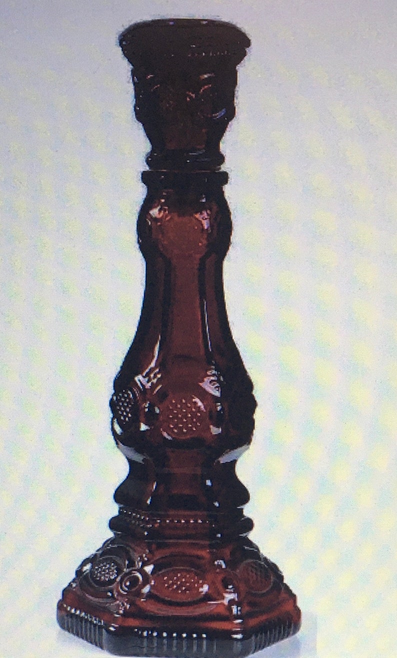 May include: A dark red glass candle holder with a decorative design. The holder has a wide base and a tall, slender stem.
