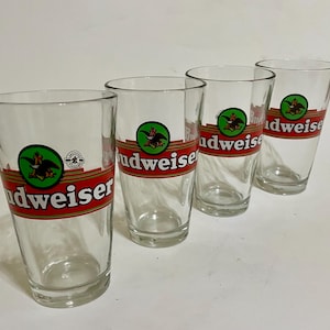 May include: Four clear glass Budweiser beer glasses with red, gold, and green logos. The glasses are empty and sitting on a white surface.