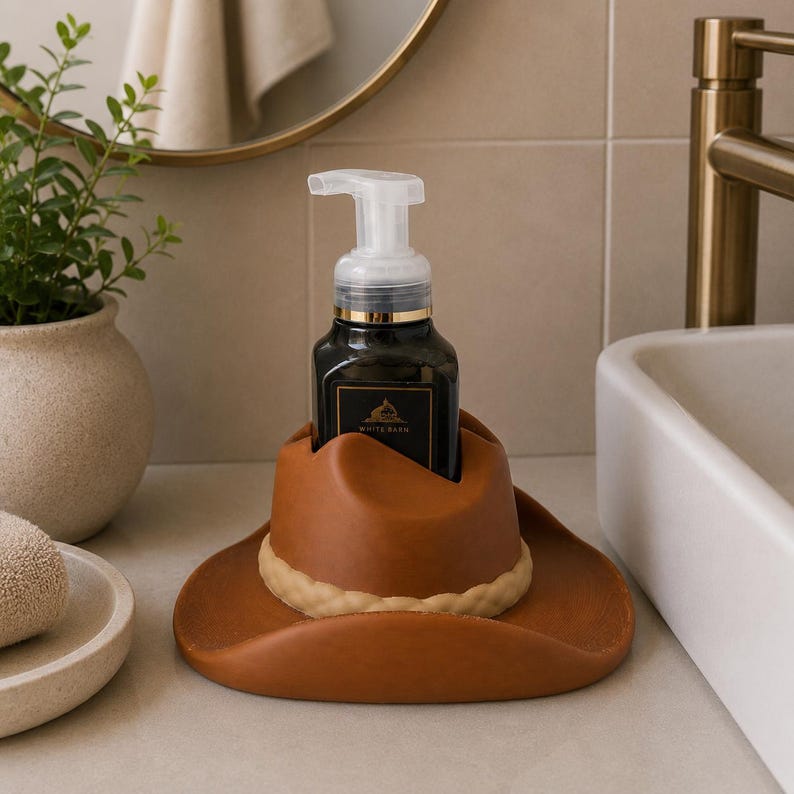 May include: A brown cowboy hat-shaped soap dispenser holder. The holder is made of a leather-like material and has a light tan band around the base of the hat. A black soap dispenser with a white pump sits inside the holder.