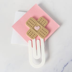 May include: A white paperclip bookmark featuring two tan adhesive bandages arranged in an X shape. A small, light pink heart is centered where the bandages overlap. The bookmark is positioned over a square, light pink piece of paper.