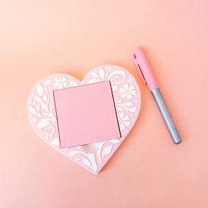 May include: A pink heart-shaped memo holder with a square opening for notes, decorated with white floral and leaf patterns. A pink pen with silver accents rests beside it on a matching pink surface.