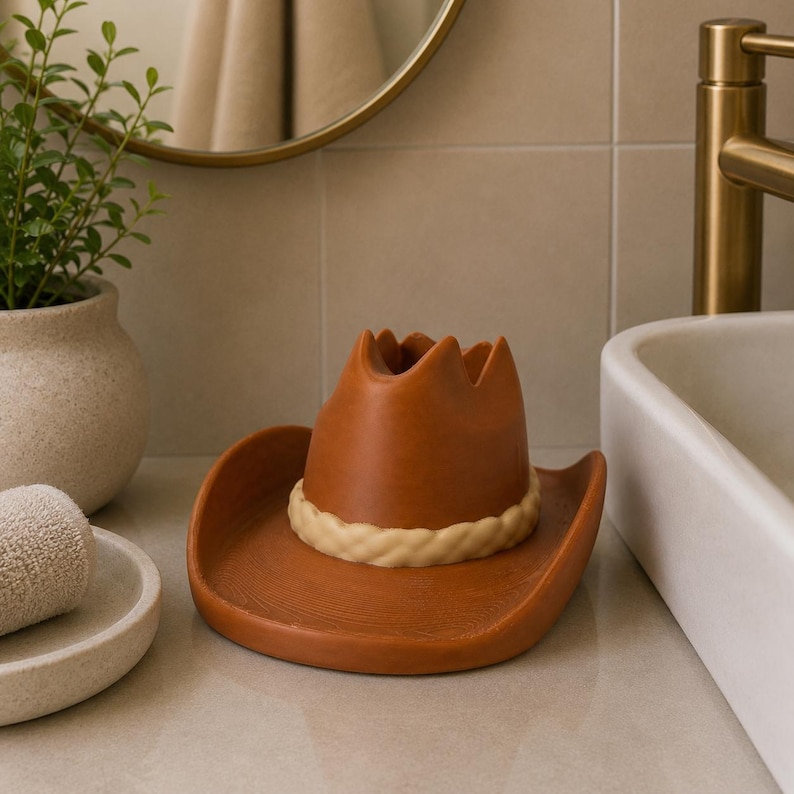 May include: A brown cowboy hat-shaped soap dish with a cream-colored rope detail. The dish is made of ceramic and sits on a bathroom counter. A small plant in a beige pot and a rolled towel are in the background.