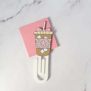 May include: A large white paperclip featuring a flat, decorative topper shaped like an iced coffee cup. The cup is tan with white ice cubes, a pink sleeve with white flower outlines, a pink bow on the lid, and a white straw extending upward.