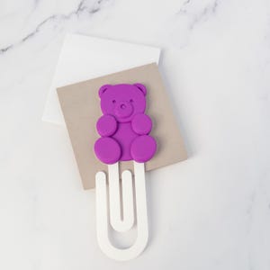 May include: A purple bear-shaped topper attached to a large white paperclip. The bear has a rounded head, small ears, and circular limbs. The paperclip is white and features a classic U-shaped design, providing a functional clip for holding papers.