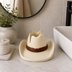 May include: A cream-colored cowboy hat-shaped decorative holder with a brown band. The hat is made of ceramic and is designed to hold small items. The holder is placed on a marble countertop in a bathroom setting.
