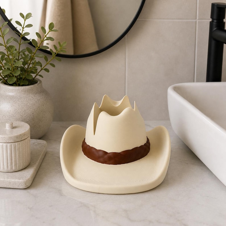 May include: A cream-colored cowboy hat-shaped decorative holder with a brown band. The hat is made of ceramic and is designed to hold small items. The holder is placed on a marble countertop in a bathroom setting.