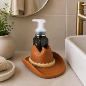 May include: A brown ceramic cowboy hat soap dispenser holder with a white rope-like trim. A black soap bottle with a white pump sits in the holder. The dispenser is on a bathroom countertop with a sink and mirror in the background.