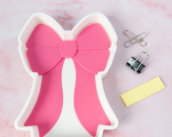 May include: A white desk organizer tray shaped like a large bow with a bright pink interior inlay. The tray features a central knot and two long, flowing ribbon tails. Beside the tray are two silver paper clips, a black binder clip, and a yellow note.