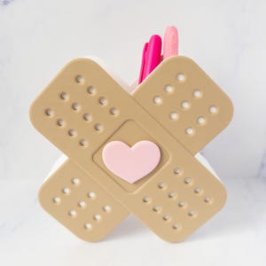 May include: A beige desk organizer shaped like two crossed adhesive bandages with a light pink heart in the center. The organizer features a grid of small circular cutouts across its surface and contains two bright pink pens inside the holder.