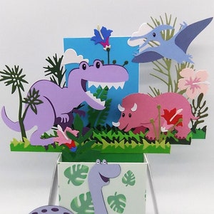 May include: A dinosaur-themed pop-up card. The card features a purple Tyrannosaurus Rex, a pink Triceratops, and a blue Pterodactyl. Green foliage and flowers surround the dinosaurs, set against a light blue backdrop. A light purple Brontosaurus is on the front.