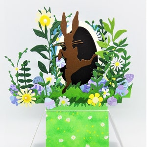 May include: A 3D pop-up card featuring a brown rabbit silhouette hopping out of an egg-shaped hole surrounded by green leaves, purple and blue flowers, and white daisies. The card is set against a green background with white daisies.