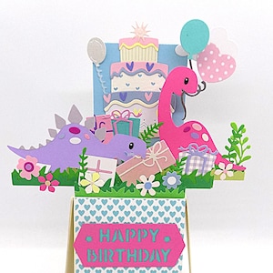 May include: A pop-up birthday card with a pink dinosaur, purple dinosaur, and a multi-layered cake. The card has a green grassy base with flowers, gifts, and balloons. The card's base is decorated with blue hearts and the words "HAPPY BIRTHDAY".