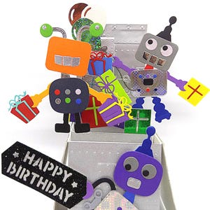 May include: A pop-up birthday card featuring colorful robot characters and gift boxes. The card has a silver metallic background and a black tag that reads "HAPPY BIRTHDAY". The robots are orange, gray, and purple with various details.
