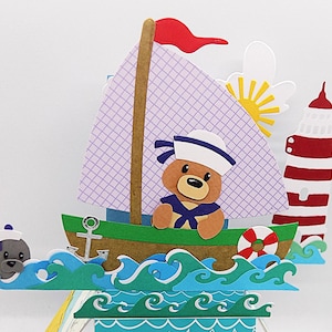 May include: A handmade paper craft featuring a teddy bear wearing a sailor hat, sailing a boat with a red and white striped lighthouse in the background. The boat is surrounded by blue and green waves.