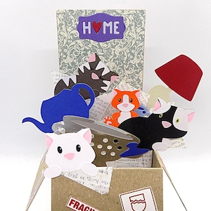 May include: A pop-up card with cartoon cats, a blue teapot, and a red shape. The card is decorated with the word "HOME" and the word "FRAGILE" on the box. The background has a floral pattern. The card is handmade.