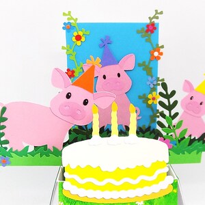 May include: A pop-up card featuring three pink pigs wearing party hats and a yellow cake with three candles. The pigs are positioned in front of a blue background adorned with green vines and flowers.