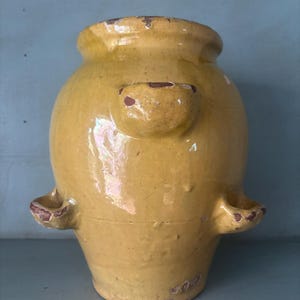 May include: A mustard yellow glazed ceramic jar with a wide mouth and three small, protruding handles spaced around the upper body. The surface features a glossy finish with visible signs of wear, including chips and exposed reddish-brown clay.