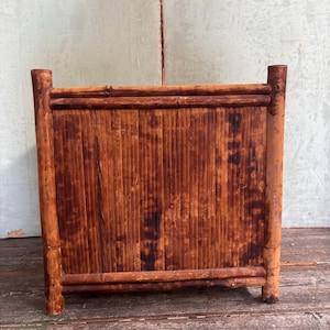 May include: A square planter box constructed from dark brown bamboo with a vertical slat design. The structure features four thick bamboo corner posts and horizontal bamboo framing along the top and bottom edges, showcasing a natural wood grain.