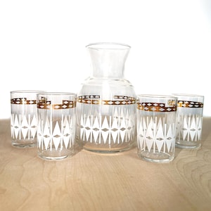 Puede incluir: Un juego de cinco jarras y vasos de vidrio transparente con un patrón geométrico blanco y dorado. La jarra y los vasos tienen una banda dorada alrededor del medio.