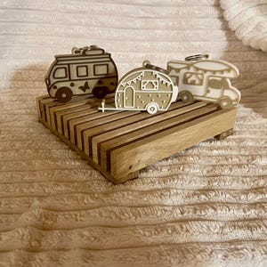 May include: Three camper-shaped keychains in beige and white, displayed on a wooden platform. The keychains feature detailed designs of RVs and trailers. The platform is made of light-colored wood with a striped pattern.