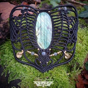 Puede incluir: Pulsera de macramé negro con una gran piedra ovalada de labradorita, cuentas doradas y detalles dorados.