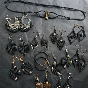 Op de afbeelding: Een assortiment zwarte macramé oorbellen en een ketting met een centraal kristal. De oorbellen hebben verschillende geometrische en bloemmotieven, sommige met zilveren of gouden accenten. De sieraden worden gepresenteerd op een donkere ondergrond, waarbij het ingewikkelde knoopwerk wordt benadrukt.