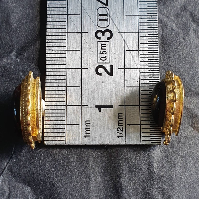 May include: Two gold-toned cufflinks with red and black gemstones. The cufflinks are measured against a ruler, showing they are approximately 1/2 inch in diameter.