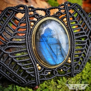 Puede incluir: Un brazalete de macramé negro con una gran piedra ovalada de labradorita azul engastada en un bisel dorado. El brazalete tiene un diseño delicado e intrincado.