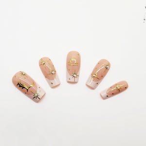 May include: Five artificial nails with a nude base, white tips, and gold star and Libra designs. The nails are arranged in a fan shape against a white background. The Outlaw Claws logo is in the upper right corner.