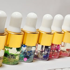 May include: A row of small glass bottles with gold caps and white droppers. The bottles contain colorful liquids and floral elements. Each bottle is labeled with a different name, such as "Queen B" and "Pistachio Pop".