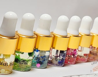 May include: A collection of small glass bottles with gold lids and white droppers. The bottles are filled with colourful liquids and floral decorations. Each bottle is labelled with a different name, such as "Queen B" and "Pistachio Pop".