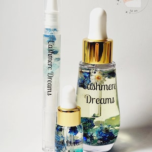 May include: Two bottles of nail oil with a clear base and gold caps. The bottles are filled with a clear liquid and have blue and white flowers inside. The bottles are labeled "Cashmere Dreams".