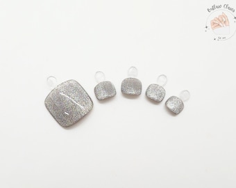 Puede incluir: Un juego de cinco uñas postizas para los pies con purpurina plateada. Las uñas son de forma cuadrada y tienen un acabado brillante y reflectante. Cada uña está adherida a una pestaña de plástico transparente. La imagen incluye un logotipo en la esquina superior derecha.