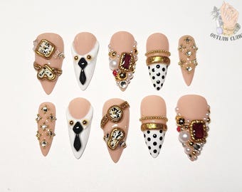 May include: A set of ten almond-shaped artificial nails with various designs. Some nails feature watch faces, gold accents, and rhinestones. Others have a black and white polka dot pattern or a white tip with a black tie design. The nails are a nude color.